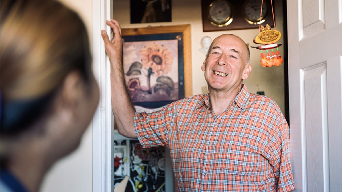A man smiling and talking to someone at the doorway of his home.