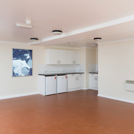 Empty communal room with a kitchenette, storage cupboards, and open floor space.