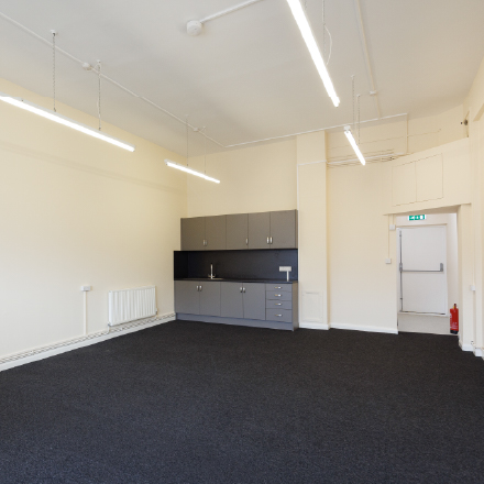Bright community room with open floor space, storage cupboards, and a kitchenette area.