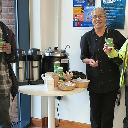 Limelight cafe worker stood around a table with tea/coffee on it. Stood with a group of visitors to the warm hub.