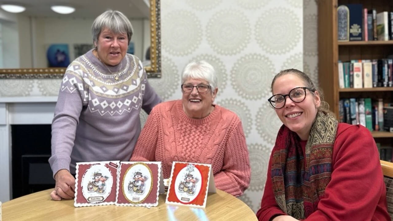 Three women smiling at a table indoors, displaying handmade greeting cards during a craft session