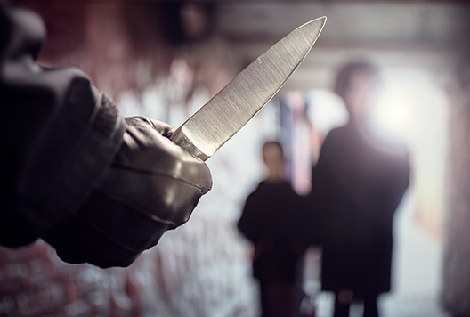 Hand in foreground holding a knife with two figures of people in the background.