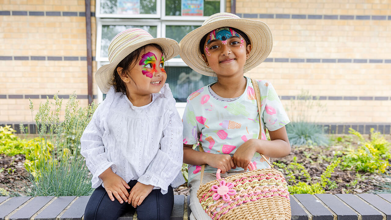 Two young girls with face paint and sun hats sit smiling on a bench in front of a garden and brick wall.