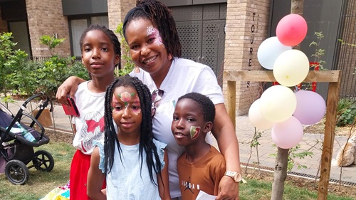 A woman with face paint, standing with three children, two of whom also have face paint. They are outdoors with balloons and greenery in the background.