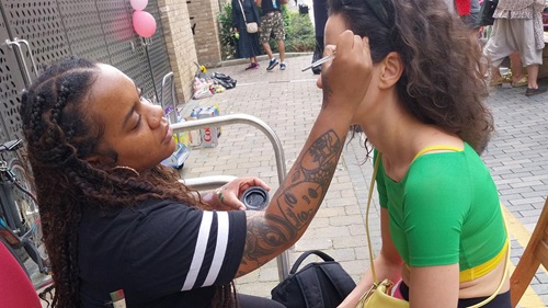 A woman with braided hair and tattoos is applying face paint to another woman. They are sitting outdoors with people in the background.