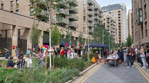 A community gathering in an urban area with people mingling, colorful banners, and apartment buildings in the background.