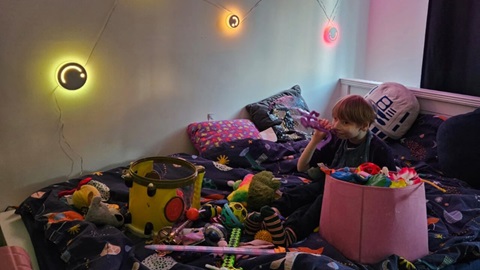 Child playing with toys on a bed in a sensory room decorated with colourful lights and soft furnishings.