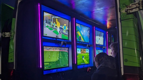 Children play video games on multiple wall-mounted screens inside a mobile gaming truck, illuminated with colourful LED lighting.