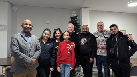 Group of adults and a young person standing together indoors in front of a decorated Christmas tree, smiling at the camera during a festive community event