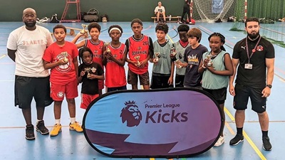 Group of youth with leaders stood behind Premiere League Kicks sign at indoor basketball event.