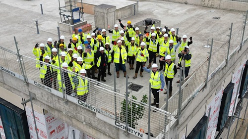 Large group of construction workers and partners in high-vis vests and hard hats gathered on the Beam Park site, waving to camera.