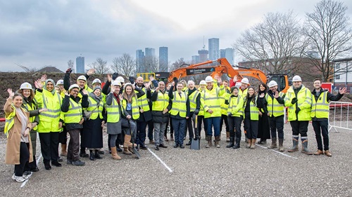 Large group of people in high‑visibility clothing gathered on a construction site in front of an excavator. Manchester high rises can be seen in the far background.