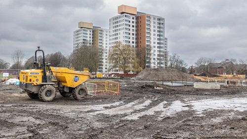 Muddy construction site with machinery, fencing and high‑rise buildings in the background.