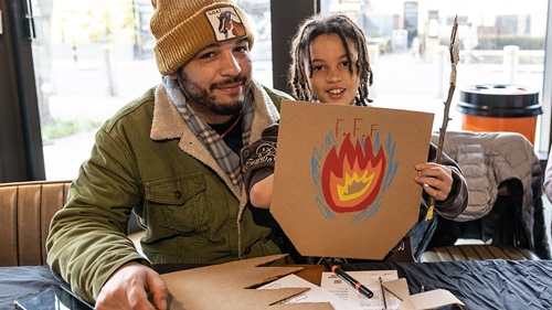 Adult in green jacket and yellow beanie hat sat smiling at the camera next to young person smiling at the camera and holding a piece of cardboard up. The cardboard has a drawing of fire on it with the letter 'F.'F.F' above the drawing.