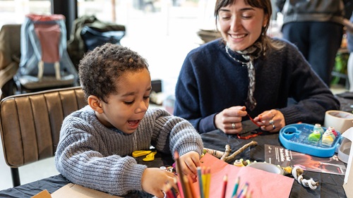 Young person with a lot of joy on their face, sat interacting with craft material around them. next to them is an adult smiling at them and holding a red thin ribbon in their hand.