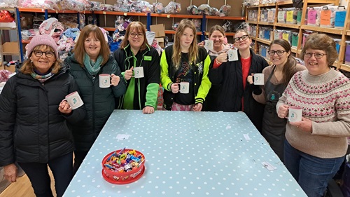 Volunteers at a community donation center enjoying a tea break, holding branded mugs, surrounded by organised supplies. 