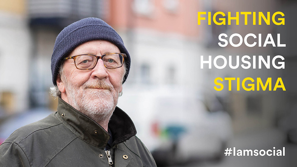 Headshot of Dave looking at the camera smiling. Dave is wearing a dark blue beanie and grey jacket with the collar popped up. On the right of the image is text that reads: FIGHTING SOCIAL HOUSING STIGMA #iamsocial