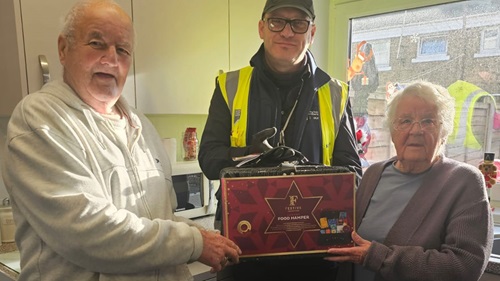 An elderly couple stands with a Durkan representative holding a festive food hamper in their kitchen.