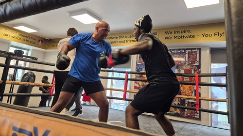 Two men sparring in a boxing ring, one in a blue shirt and the other in black, under motivational posters on the gym walls.