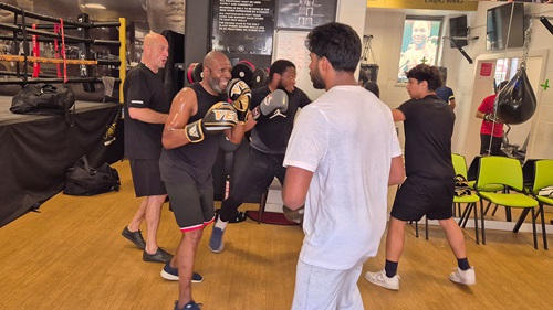 A group boxing session in a gym, with one man holding pads while others practice punches in the background.
