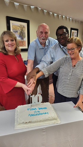 Colleagues and resident stood around cutting a white cake with 'Fiona Gardens 10yr Anniversary' written in blue on it.