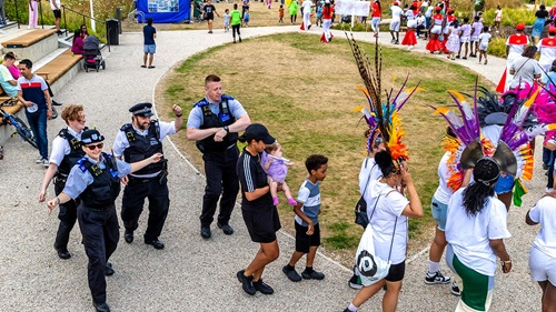 A vibrant outdoor community event featuring police officers walking alongside families and people dressed in colorful carnival costumes.
