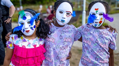 Three children posing with decorative masks adorned with feathers and painted designs at a community event.