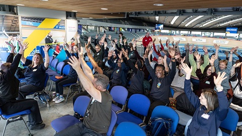 Large group of adults seated indoors raising their hands together during an activity session at a sports venue