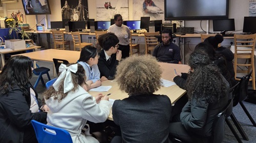 A group of students sits around a table in a classroom, engaged in discussion with two facilitators standing and sitting at the front. Computers and posters decorate the background.