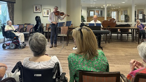 A small group of people watches a violinist and pianist performing in a cozy, well-lit room.