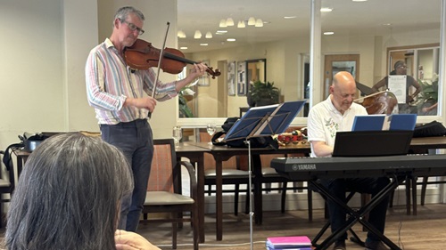A violinist plays passionately while an elderly audience listens, with a pianist accompanying him in a warm, inviting space.