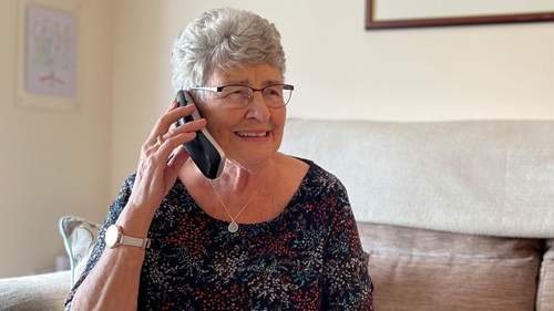 Elderly woman sat on sofa, holding a smart phone to her ear.