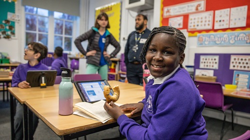 Student in classroom holding a bagel while reading a book at desk.