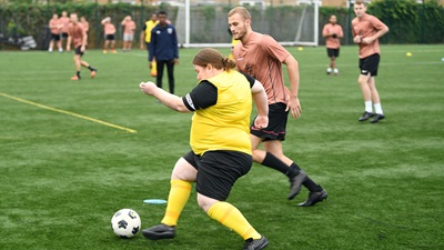 Two football teams playing a match on a grass pitch. One team is wearing salmon pink jerseys and the other have sunflower yellow jerseys. 
