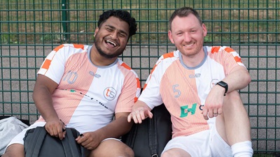 Two football team mates sat next to one another in salmon pink + white jerseys smiling at the camera.