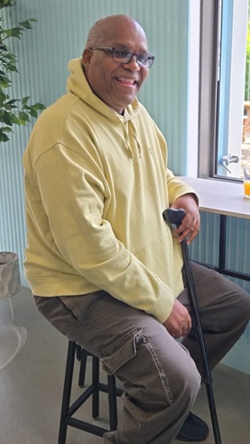 Oliver, wearing a yellow hoodie seated by window, holding a cane, indoor setting with plants.