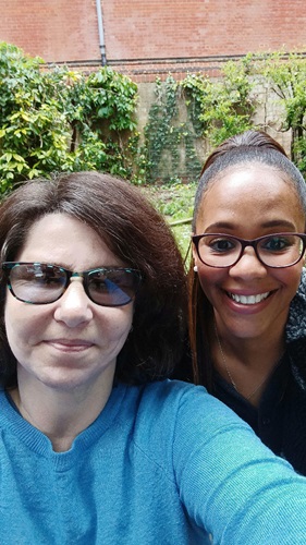Selfie outdoors with Tabitha and another person wearing glasses, greenery and brick wall in background.