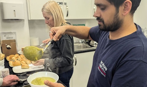 Man serving hot soup from a ladle into a bowl while another person prepares bread rolls in a community kitchen.