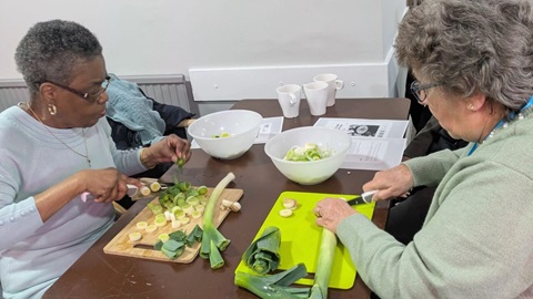 Two older women chopping leeks on cutting boards while preparing food together at a table during a cooking activity.