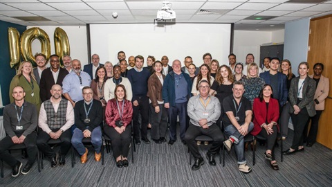 Large group of colleagues posing together indoors for a team photo, with gold “100” balloons displayed behind them.