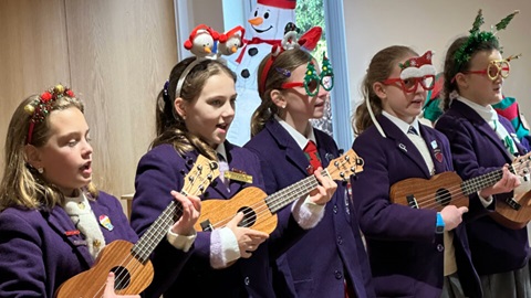 Children wearing festive headbands and glasses play ukuleles and sing during a Christmas performance indoors.