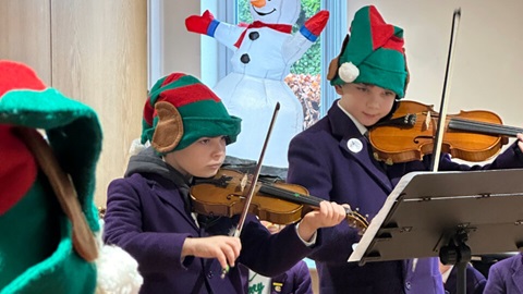 Two children in elf hats play violins from sheet music during a festive indoor performance, with a snowman decoration behind them.