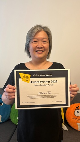Adeline holding an award certificate indoors, which says, "Volunteers Week Award Winner 2026 – Open Category” - She is in front of a sofa with colourful M&M cushions on it..