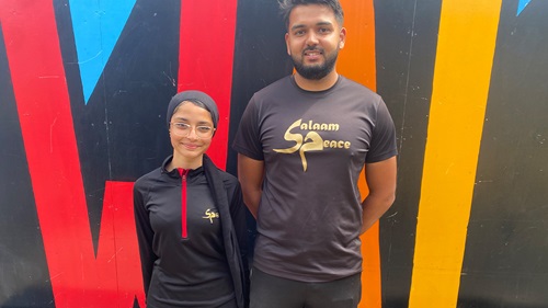 Mumtaz and Azi wearing Salaam Peace shirts, standing against colorful wall.