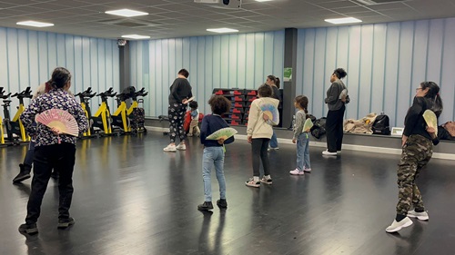 A mixed age group of people facing away from the camera at a dance class. They are holding paper fans behind their back as they dance.