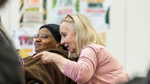 Two people sitting close together, one leaning in to speak during a community event with posters in the background.