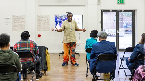 Person speaking to a seated audience in a community hall with presentation screen and posters on the wall.