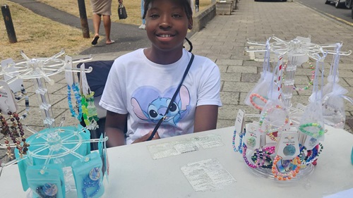 Young person sitting at a street table with colorful beaded jewelry on display racks. They wear a shirt with a cartoon character and smile warmly.