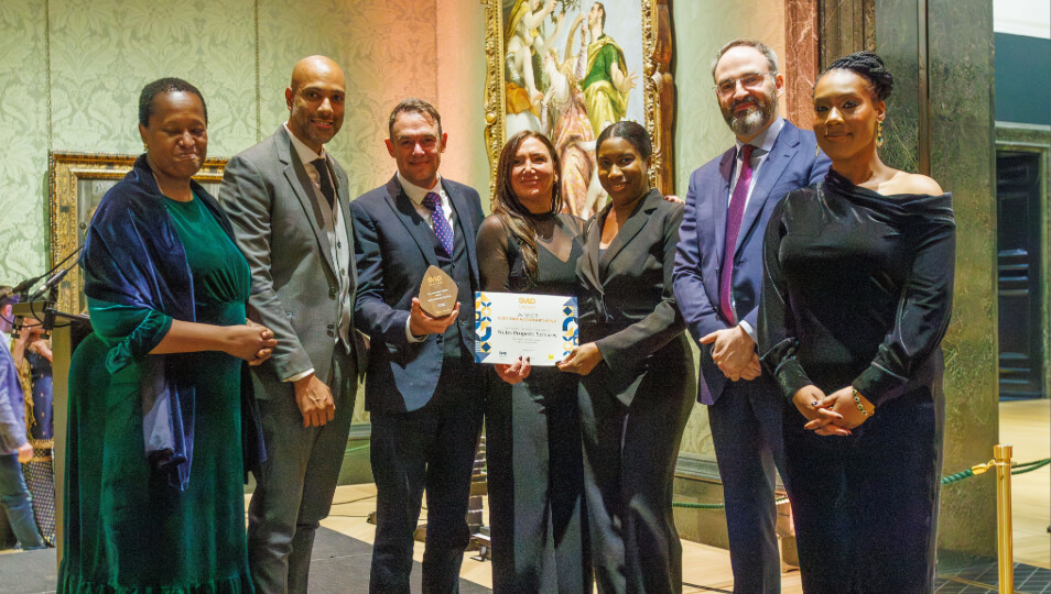 Group of people in formal attire stand together at an awards event, smiling and holding a certificate and trophy inside an ornate venue.
