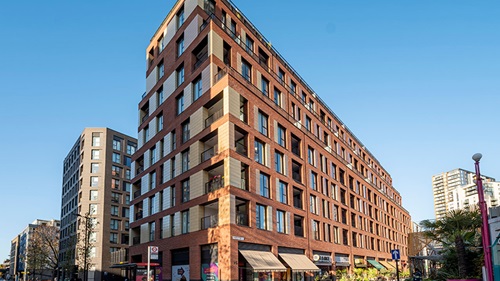 Modern red brick building with balconies, large windows, shops below, and a clear blue sky above